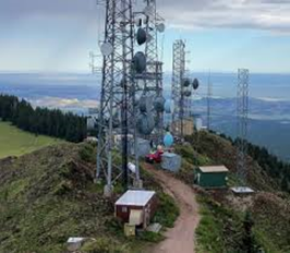 Telecommunications towers with multiple dish antennas on a mountain ridge, with a dirt access road and small utility buildings overlooking a distant valley.